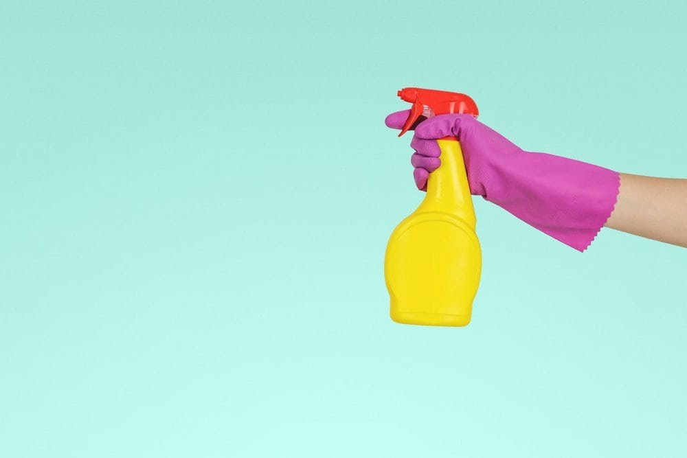 A hand wearing a pink cleaning glove holding a yellow spray bottle.