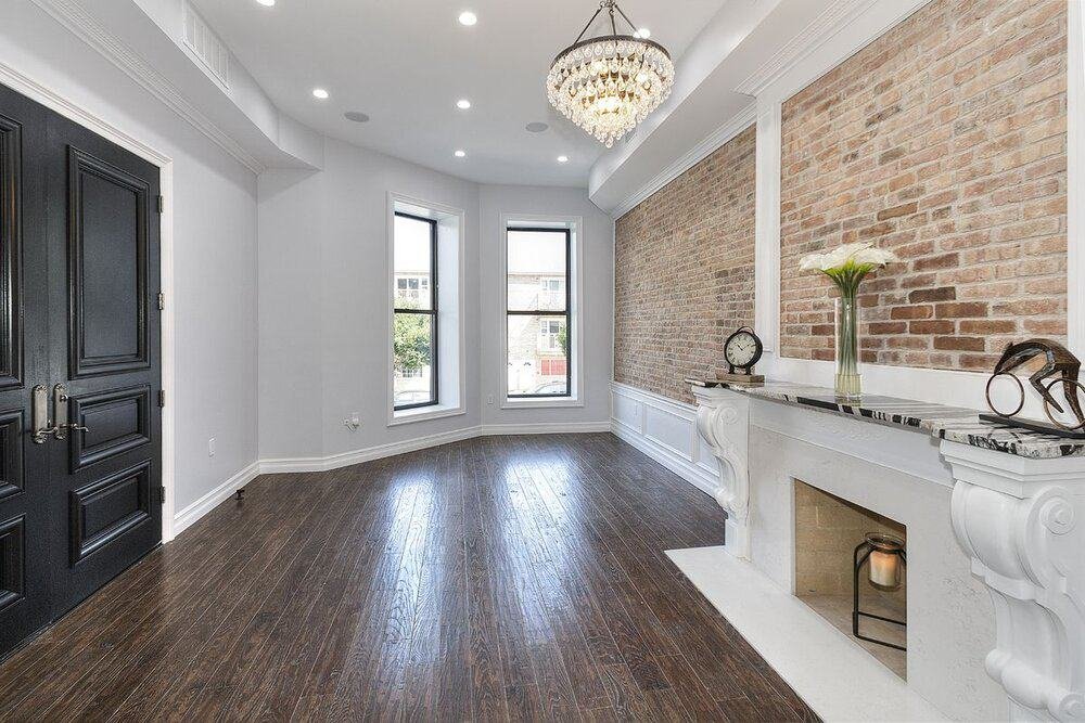 Elegant living room featuring polished dark wood floors, light gray walls, and a white ceiling with recessed lighting. The room includes large windows, a black door, and an ornate white fireplace with a brick wall above it. A crystal chandelier hangs in the center, adding a touch of luxury
