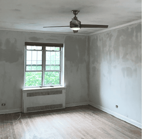 Photo of a room in the process of renovation with unfinished, patchy walls and a bare hardwood floor. There is a window and a ceiling fan present in the room.