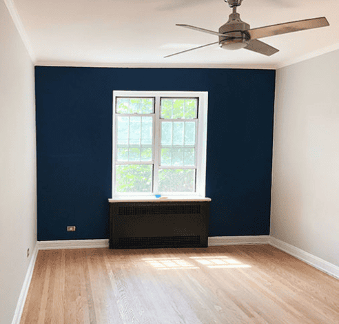 Photo of a room with one wall painted in a deep blue, contrasted by surrounding white walls. The room features a large window, hardwood floors, and a ceiling fan.