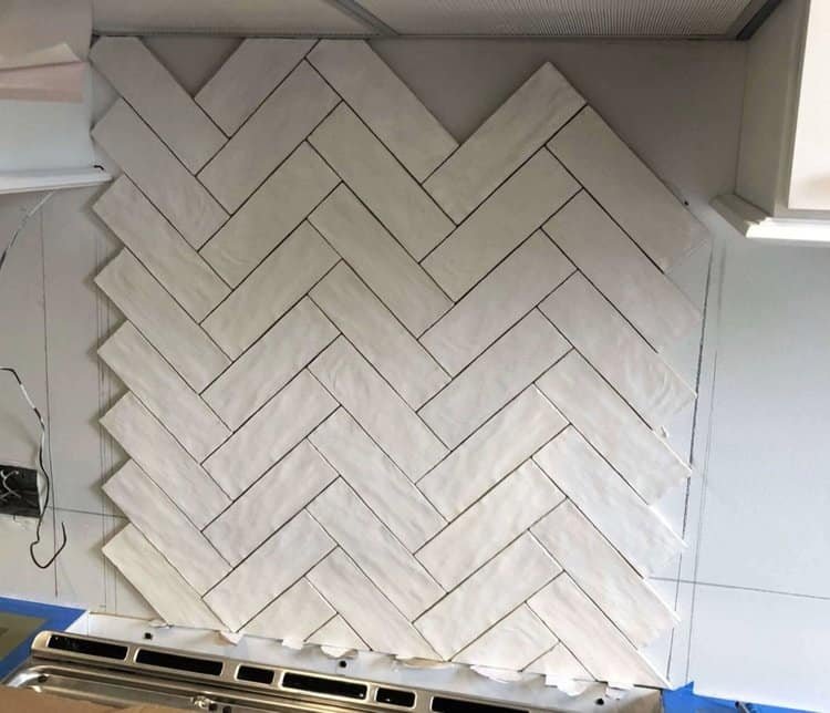 A kitchen backsplash in progress, featuring white tiles arranged in a herringbone pattern above a stove area.