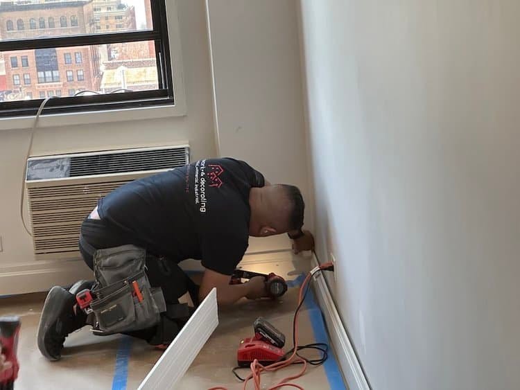 A worker installing baseboards using a power drill in a room with taped-off floors and walls.