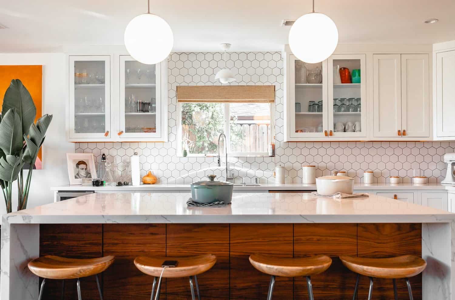 Photo of a bright, modern kitchen featuring freshly painted white cabinets with glass fronts. The cabinets contrast beautifully with the hexagonal tile backsplash and marble countertops. A large kitchen island with wooden stools and decorative items completes the stylish look.