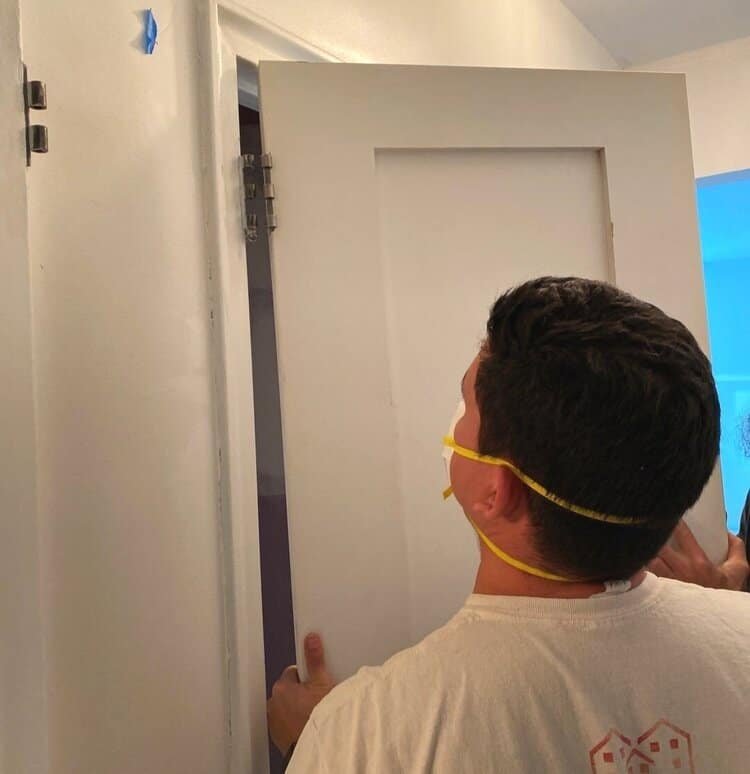 A worker wearing a mask and installing a white door in a doorway.