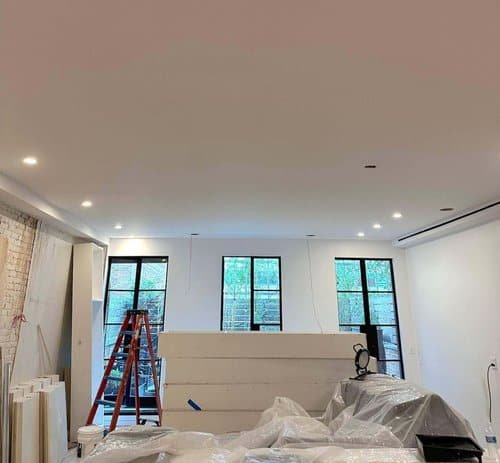 A room under renovation with drywall installed, recessed lighting, and plastic-covered furniture, with a red ladder in the background.