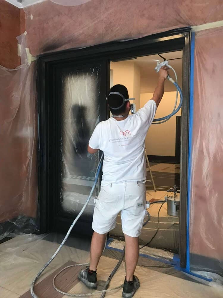 A professional painter applies electrostatic paint to a window frame using specialized equipment, while the surrounding area is protected with plastic sheeting.