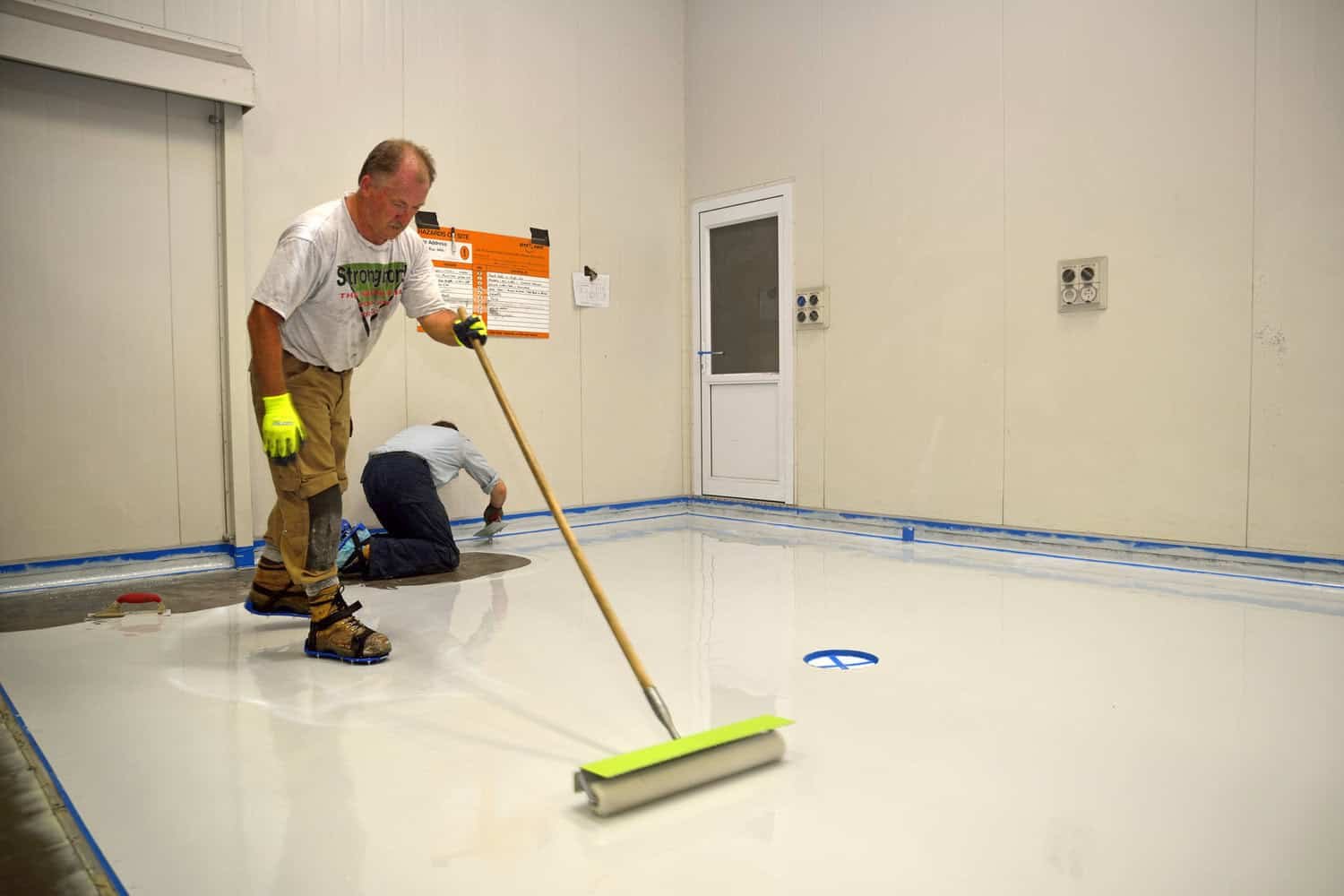 Two workers applying a white epoxy coating to a floor, one using a roller and the other kneeling to ensure an even application.