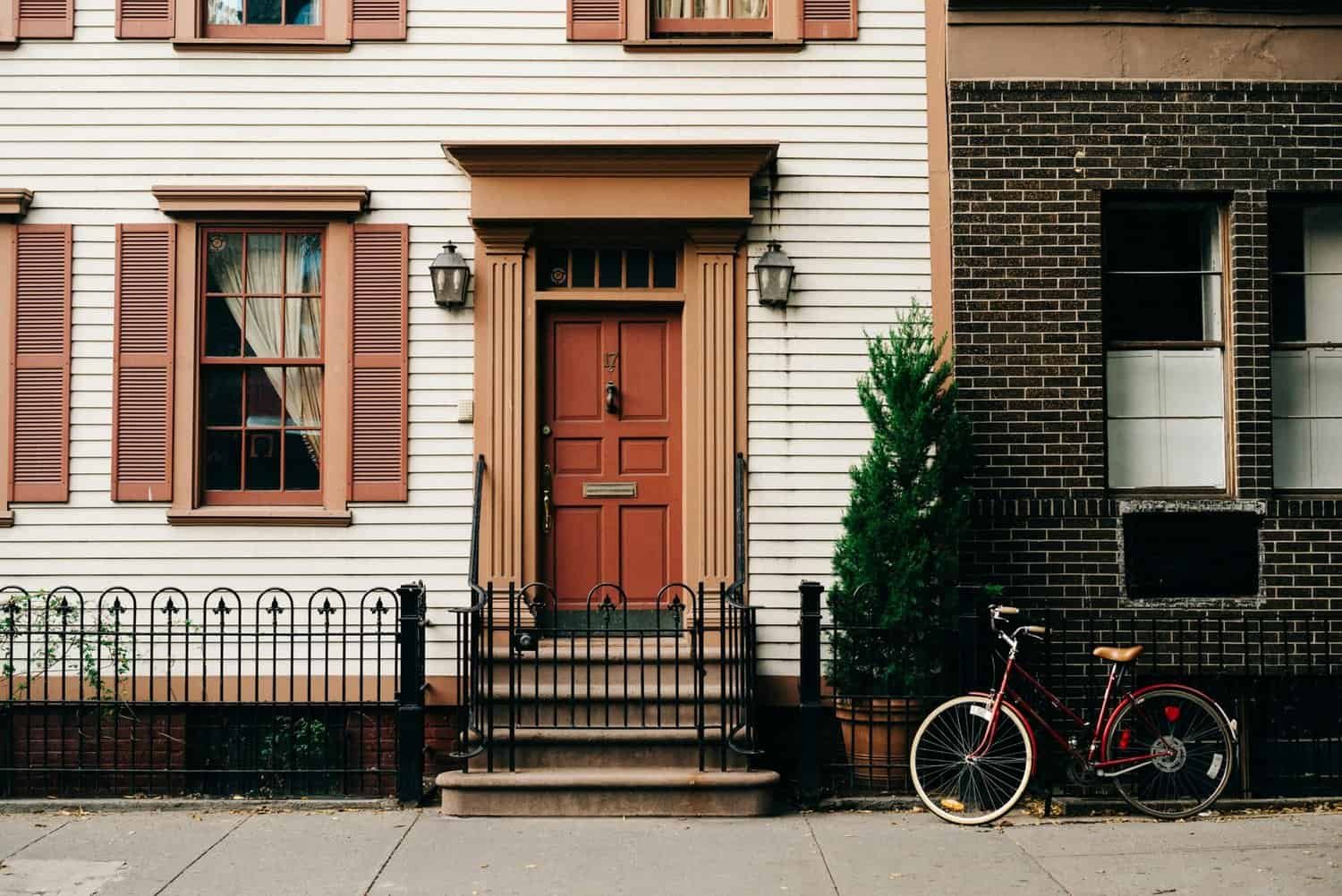 Photo of a charming house exterior featuring freshly painted white siding with red trim around the windows and door. The red front door and shutters add a vibrant touch, complemented by black wrought iron fencing and a red bicycle parked nearby.