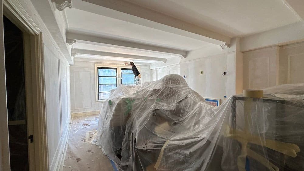 A living room covered in plastic sheets during renovation with a worker painting the ceiling.