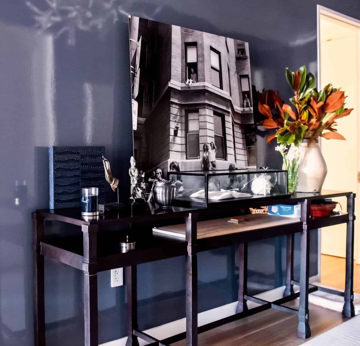 Console table against a dark glossy blue wall, decorated with a large black-and-white photo and various ornaments, showcasing a stylish, contemporary interior.