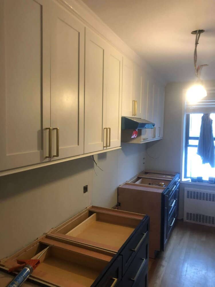 A kitchen during renovation, showing white upper cabinets with brass handles and partially installed navy blue lower cabinets.