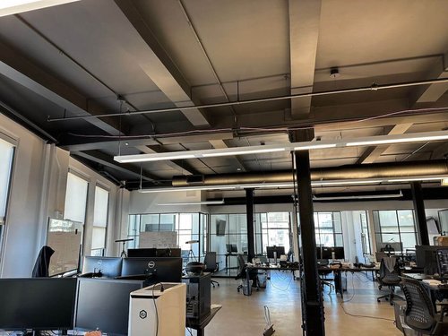 An open-plan office with exposed ceilings painted in dark tones, white walls, and numerous workstations, highlighting a contemporary and industrial design.