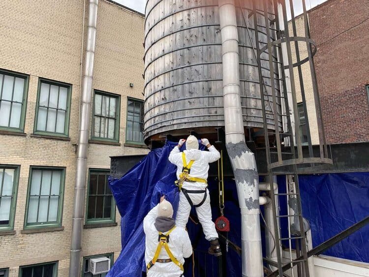 Two workers in protective white suits and yellow safety harnesses work on a large, cylindrical water tank, preparing it for painting. A blue tarp is draped around the base of the tank.