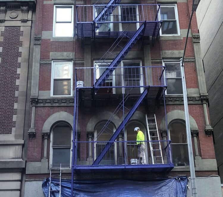 A worker in a high-visibility yellow jacket and white pants paints the metal framework of a fire escape on an older brick building. The fire escape is being painted blue, and a tarp is set up below.