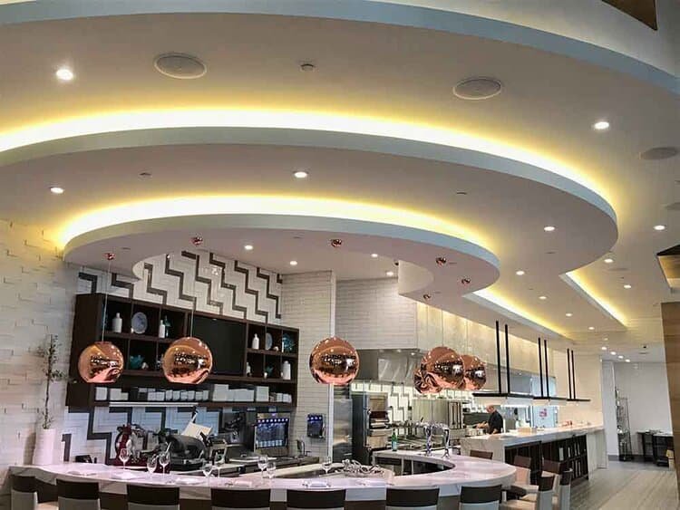A modern restaurant interior with a stylish, curved ceiling featuring integrated lighting and hanging copper pendant lights above a sleek, open kitchen.