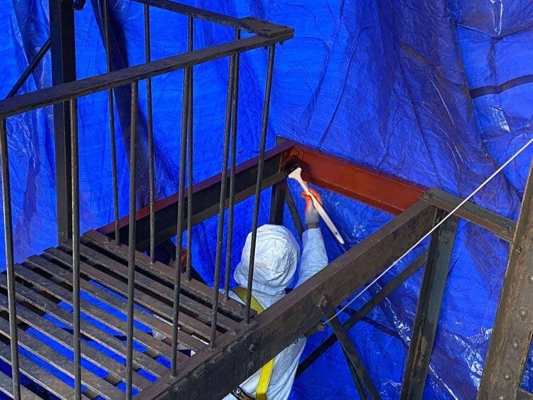 A worker in a white protective suit painting a steel structure with red primer, using a brush inside a blue-tarped area.