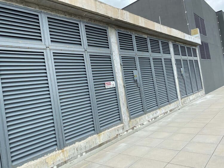 A building with gray stucco and metal louver panels, showing wear and exposed concrete at the base.