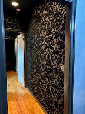 Narrow hallway with polished hardwood floors, featuring a dark, intricate floral wallpaper on one side and a light-colored door at the end.