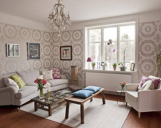 Cozy living room with light wooden floors and cream walls adorned with large patterned wallpaper. The room is furnished with white sofas, a glass coffee table, and accented with colorful cushions and fresh flowers, under a classic chandelier.