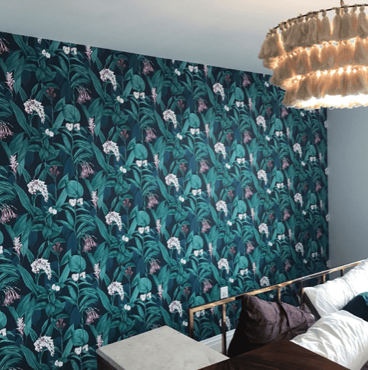 Modern bedroom wall decorated with vibrant dark blue wallpaper featuring a dense tropical leaf and flower pattern, paired with a minimalist white side table and a plush velvet cushion.